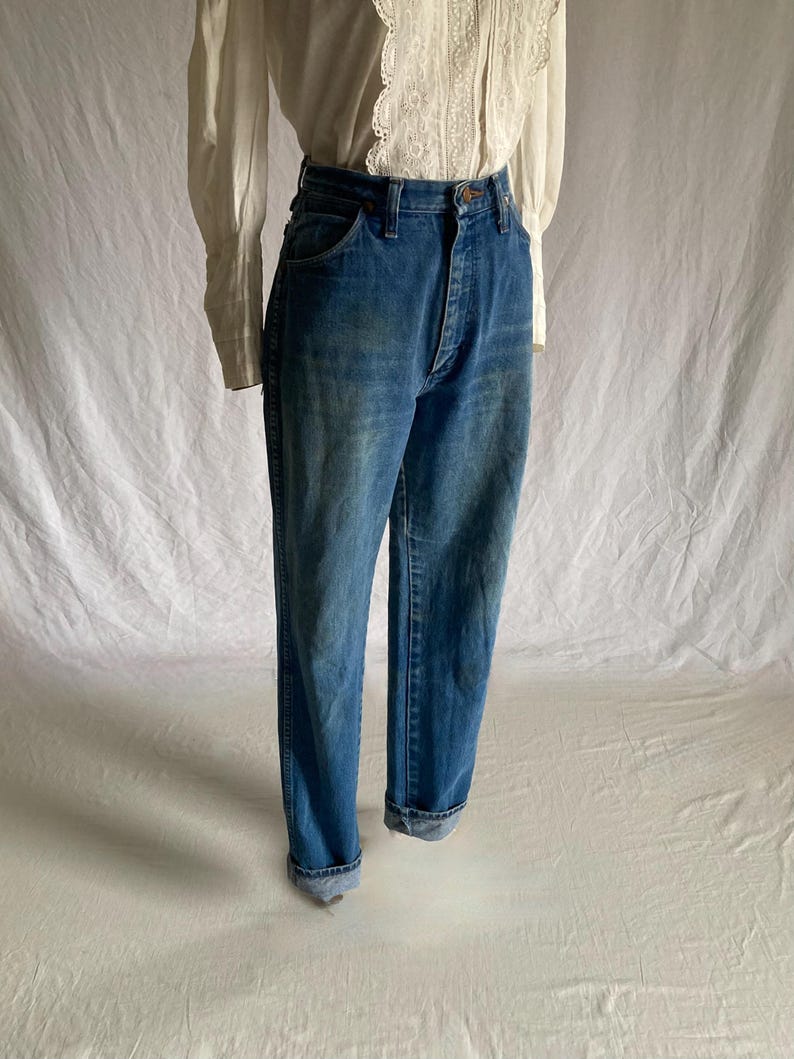 Pu&ograve; includere: Jeans blu in denim con vita alta e chiusura con bottoni. I jeans hanno un classico design a cinque tasche e sono risvoltati alle caviglie. I jeans sono abbinati a una camicetta bianca con dettagli in pizzo.