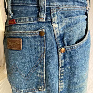 Pu&ograve; includere: Primo piano di jeans vintage in denim blu. Presenta una toppa in pelle marrone con il logo "Wrangler", rivetti in rame e un'etichetta rossa "Wrangler". I jeans hanno un classico design a cinque tasche e un lavaggio sbiadito.