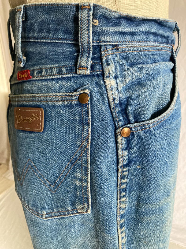 Pu&ograve; includere: Primo piano di jeans vintage in denim blu. Presenta una toppa in pelle marrone con il logo "Wrangler", rivetti in rame e un'etichetta rossa "Wrangler". I jeans hanno un classico design a cinque tasche e un lavaggio sbiadito.