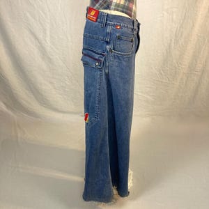 K&ouml;nnte beinhalten: Hellblaue Jeans mit weitem Bein. Die Jeans hat mehrere Taschen, darunter Cargotaschen mit roten N&auml;hten. Ein rotes Patch mit gelbem Text ist am Bund sichtbar. Die Jeans hat einen ausgefransten Saum.