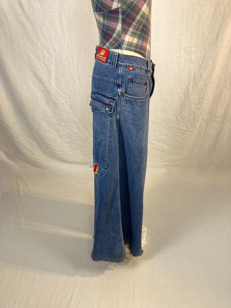 K&ouml;nnte beinhalten: Hellblaue Jeans mit weitem Bein. Die Jeans hat mehrere Taschen, darunter Cargotaschen mit roten N&auml;hten. Ein rotes Patch mit gelbem Text ist am Bund sichtbar. Die Jeans hat einen ausgefransten Saum.