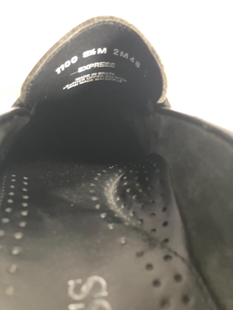 May include: Close-up of the inside of a black shoe. The shoe's interior features a perforated insole and the text "1100 8&frac12; M EXPRESS 2M48". The shoe is made in Brazil with leather upper and man-made materials.
