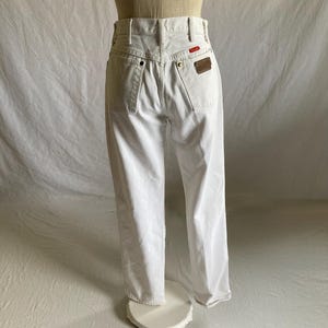 May include: White denim jeans with a classic five-pocket design. Features a red and brown leather patch on the back. The jeans are displayed on a mannequin.