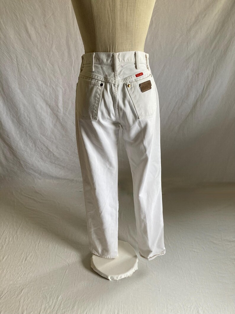 May include: White denim jeans with a classic five-pocket design. Features a red and brown leather patch on the back. The jeans are displayed on a mannequin.