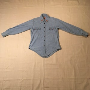 May include: A light blue denim shirt with long sleeves and a button-down front. The shirt has embroidered flowers on the collar and pockets.