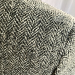 May include: A close-up view of a gray and black wool-blend blazer featuring a prominent herringbone weave pattern. The fabric has a textured, fuzzy appearance with visible fibers throughout the weave, showcasing the detailed construction of the garment.