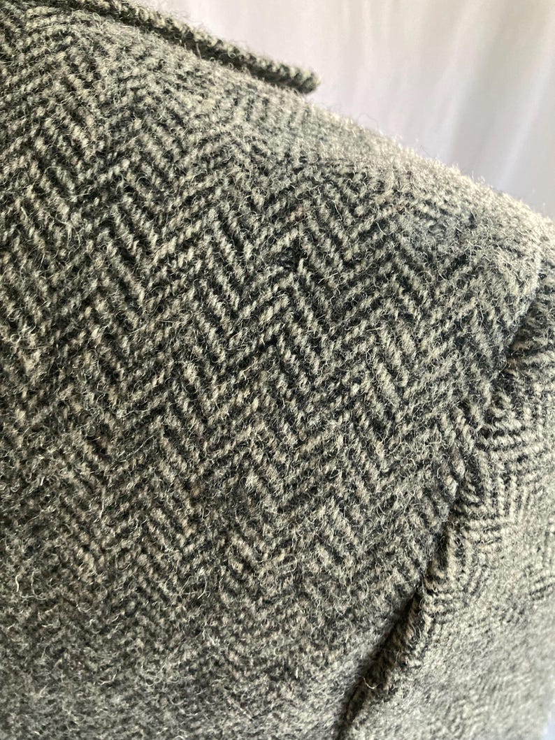 May include: A close-up view of a gray and black wool-blend blazer featuring a prominent herringbone weave pattern. The fabric has a textured, fuzzy appearance with visible fibers throughout the weave, showcasing the detailed construction of the garment.
