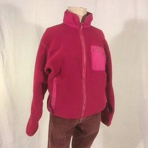 Peut inclure: Une veste en polaire rose avec une fermeture éclair et une poche rose sur la poitrine gauche. La veste a un col montant et des manches longues.