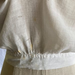 May include: Close-up of a cream-colored linen top with a gathered waist and a wide, contrasting band. The fabric shows subtle texture and slight discoloration, suggesting age or wear. The garment is displayed on a mannequin.