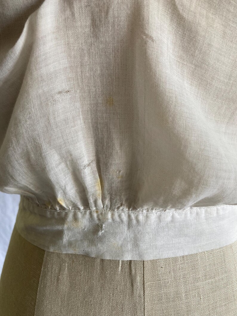 May include: Close-up of a cream-colored linen top with a gathered waist and a wide, contrasting band. The fabric shows subtle texture and slight discoloration, suggesting age or wear. The garment is displayed on a mannequin.