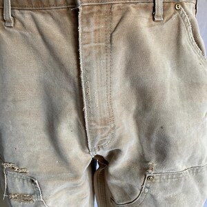 May include: Close-up of tan work pants with visible wear and tear. The pants feature multiple pockets, reinforced knees, and a button closure with the brand name. The fabric appears to be durable canvas.