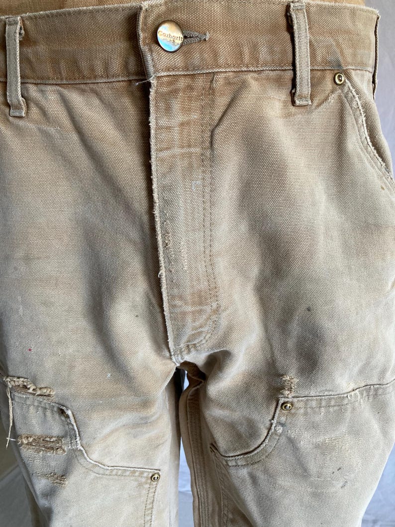 May include: Close-up of tan work pants with visible wear and tear. The pants feature multiple pockets, reinforced knees, and a button closure with the brand name. The fabric appears to be durable canvas.