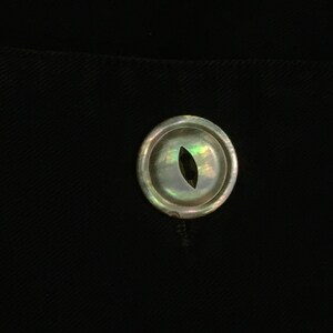 May include: A close-up of a single, round, iridescent button with a small, oval-shaped hole in the center. The button is a light, pearly white color with hints of green and blue.