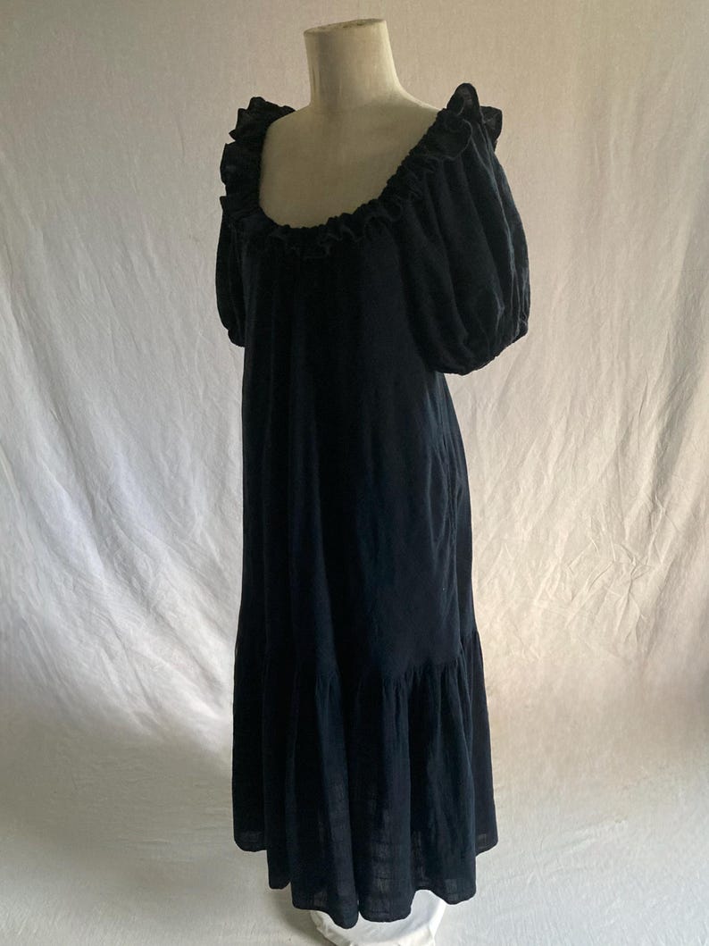 May include: A dark navy blue maxi dress displayed on a mannequin. The dress features a ruffled neckline, short puffed sleeves, and a tiered skirt. The fabric appears to be a lightweight material, suitable for warmer weather. The dress is floor-length.