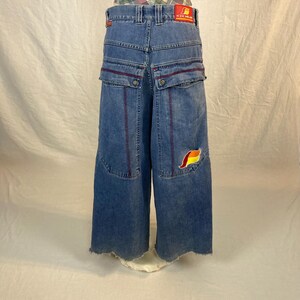 K&ouml;nnte beinhalten: Blaue Jeans mit weitem Bein, roten Akzenten und einem bunten Aufn&auml;her. Die Jeans hat gro&szlig;e Taschen und einen rohen Saum. Ein rotes Etikett mit wei&szlig;em Text ist auf der R&uuml;ckseite sichtbar.