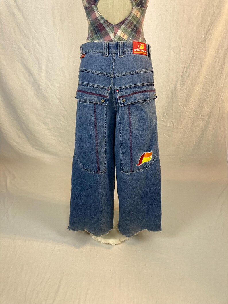 K&ouml;nnte beinhalten: Blaue Jeans mit weitem Bein, roten Akzenten und einem bunten Aufn&auml;her. Die Jeans hat gro&szlig;e Taschen und einen rohen Saum. Ein rotes Etikett mit wei&szlig;em Text ist auf der R&uuml;ckseite sichtbar.