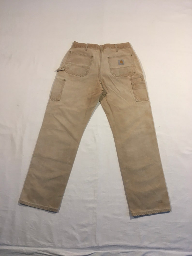 Puede incluir: Un par de pantalones de trabajo de mezclilla beige con un parche marr&oacute;n en el bolsillo trasero derecho.