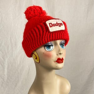Op de afbeelding: Een felrode gebreide muts met een bijpassende pompon. De hoed heeft een witte patch met het woord "Dodge" in zwarte letters. De muts wordt getoond op een mannequinhoofd.