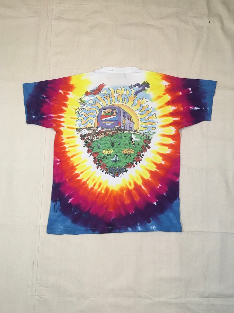 May include: Tie-dye t-shirt with a colorful swirl pattern. The shirt features a graphic of a psychedelic bus with the text "Summer Tour 1994" on the front.