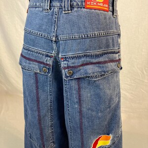 K&ouml;nnte beinhalten: Blaue Jeans mit mehreren Taschen und roten Nahtdetails. Verf&uuml;gt &uuml;ber einen roten Aufn&auml;her mit dem Text "KIK NEAR" am hinteren Bund und ein farbenfrohes Logo auf der Tasche. Die Jeans haben ein weites Bein-Design.