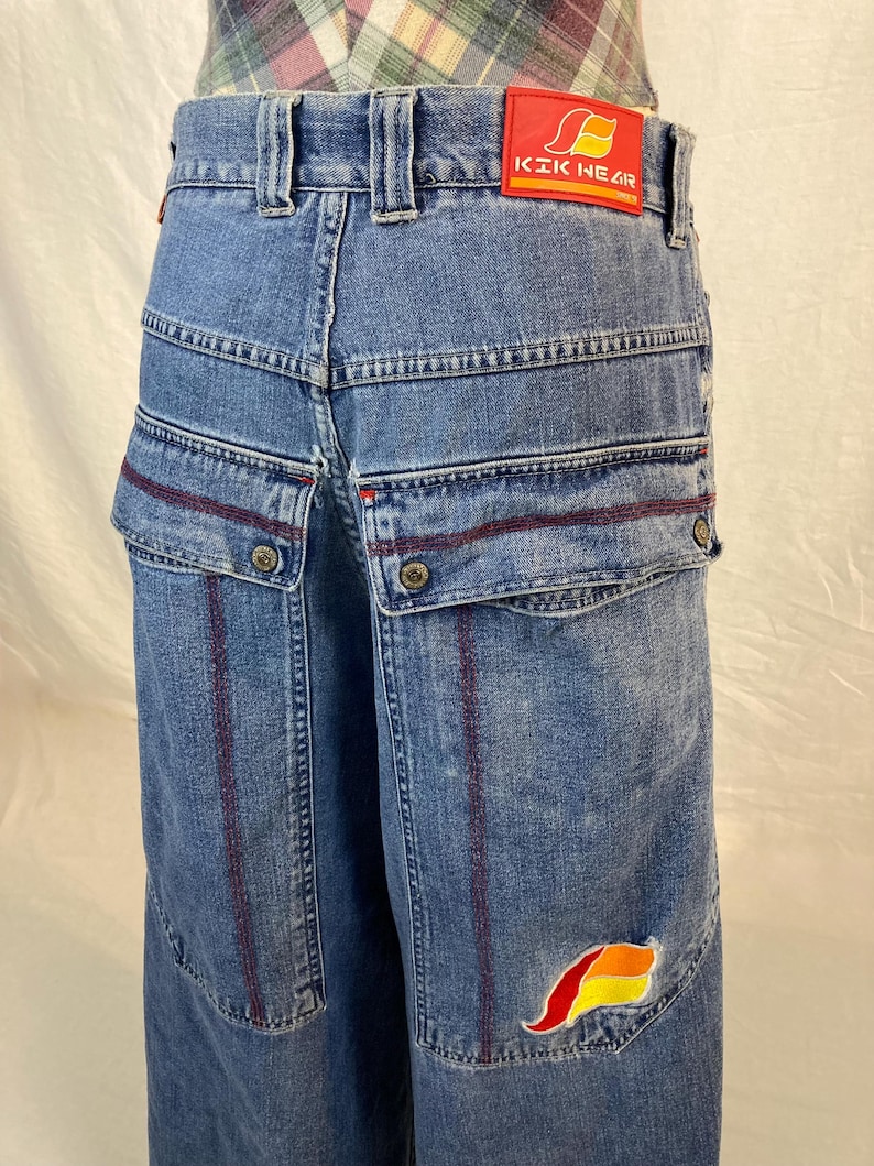 K&ouml;nnte beinhalten: Blaue Jeans mit mehreren Taschen und roten Nahtdetails. Verf&uuml;gt &uuml;ber einen roten Aufn&auml;her mit dem Text "KIK NEAR" am hinteren Bund und ein farbenfrohes Logo auf der Tasche. Die Jeans haben ein weites Bein-Design.