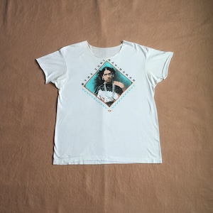 Könnte beinhalten: Weißes T-Shirt mit einem rautenförmigen Grafik, das ein Porträt einer Frau mit dem Text "The Zuni Man-Woman" um die Grafik herum zeigt.