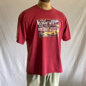 May include: A red t-shirt with a graphic print featuring a colorful scene with figures and the text "DAY OF THE GODS • GAUGUIN." The shirt is displayed on a mannequin, paired with light green pants.