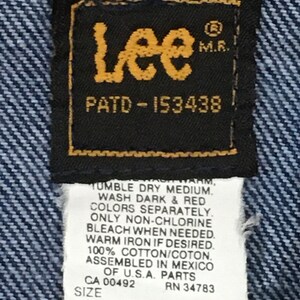 Op de afbeelding: Een blauw denim jean label met het Lee logo, de tekst "PATD-153438" en wasinstructies. Het label bevat ook de maat "46 REG."