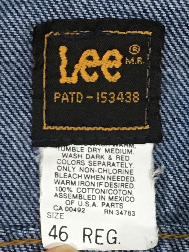 Op de afbeelding: Een blauw denim jean label met het Lee logo, de tekst "PATD-153438" en wasinstructies. Het label bevat ook de maat "46 REG."