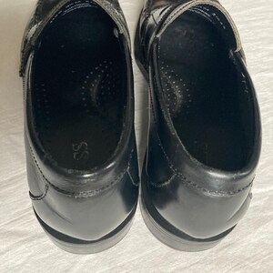 May include: A pair of black leather penny loafers. These classic shoes feature a rounded toe, a strap across the vamp, and a low heel. The interior is black with a cushioned insole. The shoes are displayed on a white surface.