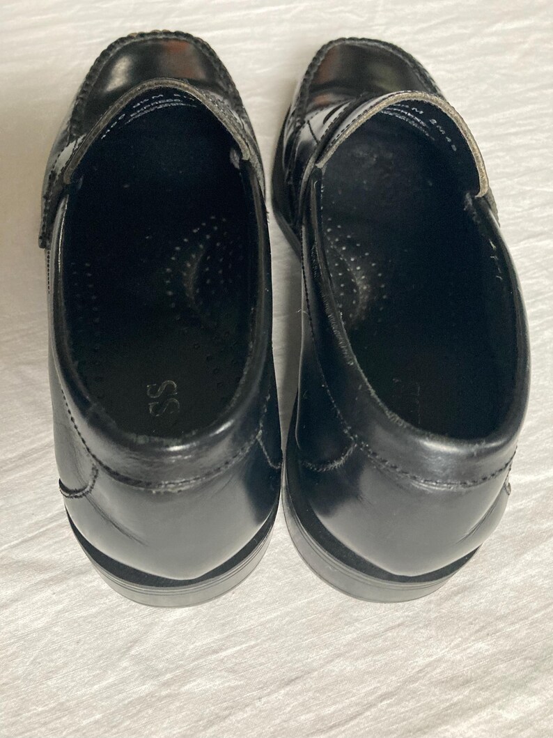 May include: A pair of black leather penny loafers. These classic shoes feature a rounded toe, a strap across the vamp, and a low heel. The interior is black with a cushioned insole. The shoes are displayed on a white surface.