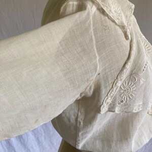 May include: A vintage white blouse with long sleeves and a decorative lace collar. The collar features intricate floral embroidery. The blouse is made of a lightweight fabric and has a slightly off-white hue, suggesting age or wear.