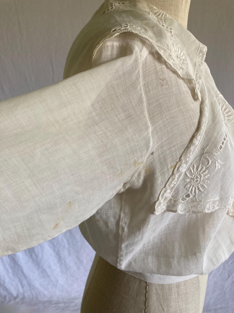 May include: A vintage white blouse with long sleeves and a decorative lace collar. The collar features intricate floral embroidery. The blouse is made of a lightweight fabric and has a slightly off-white hue, suggesting age or wear.