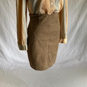 May include: A brown suede mini skirt with a high waist and geometric stitching. The skirt is paired with a button-down shirt with vertical stripes in shades of beige, tan, and light blue. The skirt appears to be vintage.