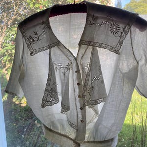 May include: A vintage white blouse with a V-neck and short sleeves. The blouse features intricate floral embroidery and lace detailing around the collar and front panels. It has small, round buttons down the front and a gathered waistline. The blouse is hanging on a black hanger.