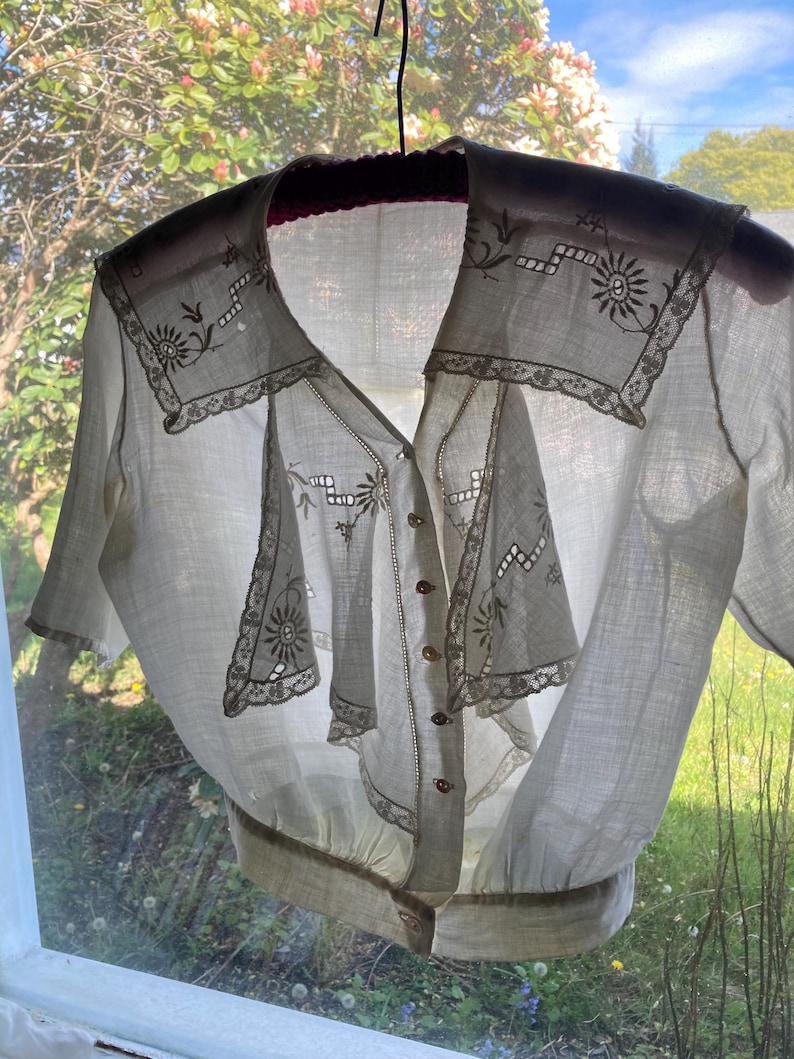 May include: A vintage white blouse with a V-neck and short sleeves. The blouse features intricate floral embroidery and lace detailing around the collar and front panels. It has small, round buttons down the front and a gathered waistline. The blouse is hanging on a black hanger.