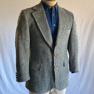 May include: A grey and white herringbone blazer featuring notched lapels, two front buttons, and two flap pockets. The blazer is worn over a dark blue button-down shirt and light-colored trousers, showcasing a structured fit and classic tailoring.