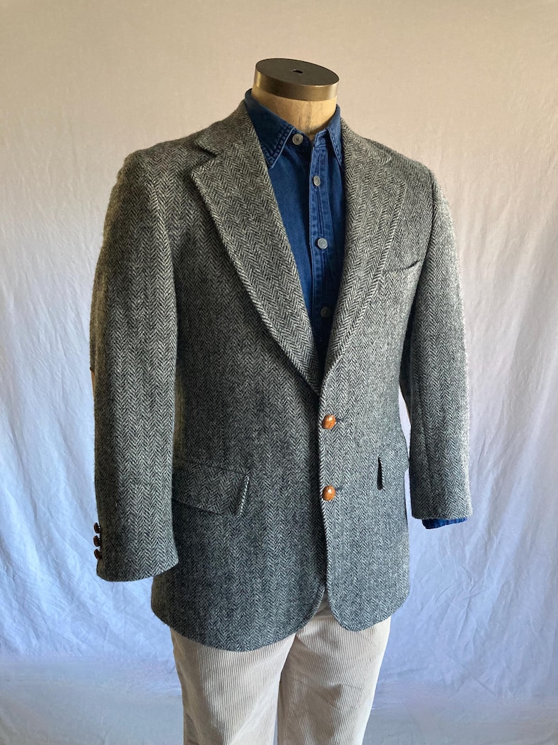 May include: A grey and white herringbone blazer featuring notched lapels, two front buttons, and two flap pockets. The blazer is worn over a dark blue button-down shirt and light-colored trousers, showcasing a structured fit and classic tailoring.