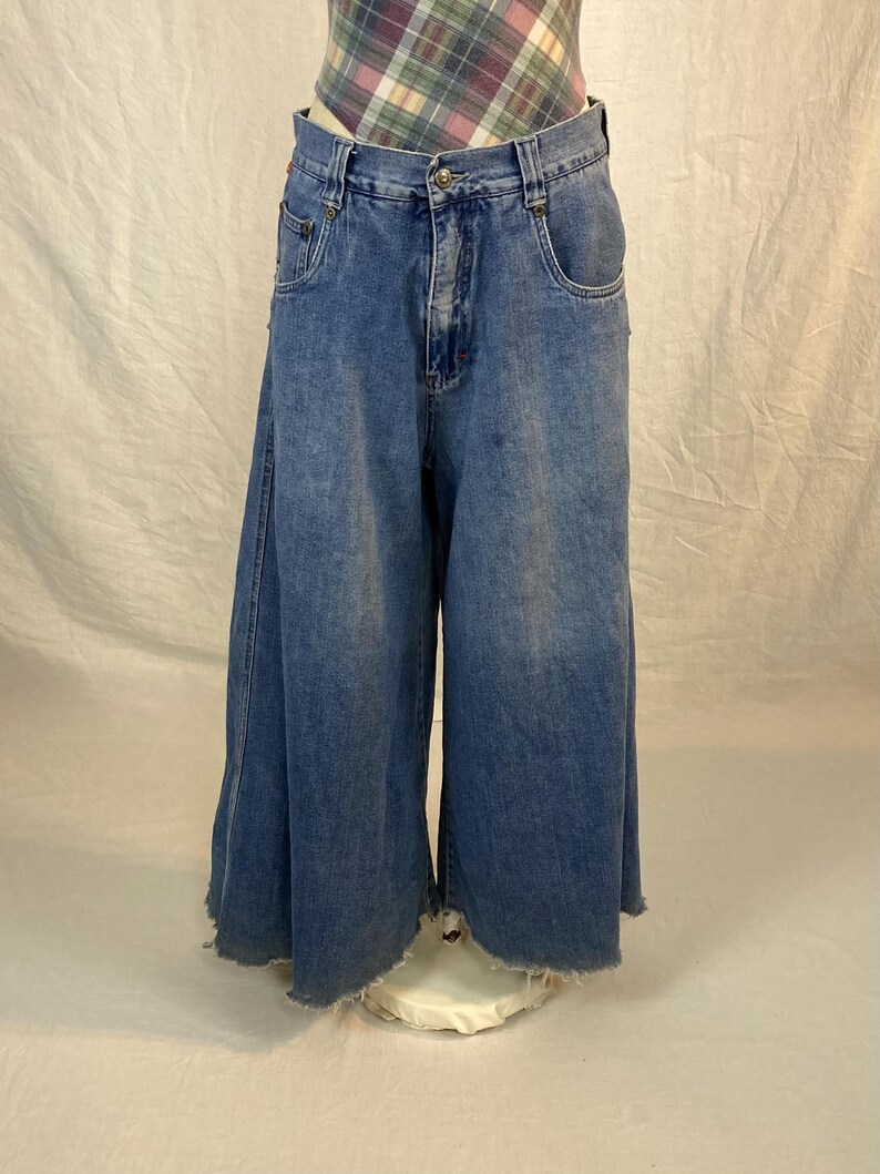 K&ouml;nnte beinhalten: Blaue Jeans-Culottes mit weitem Bein und ausgefranstem Saum. Die Jeans haben ein klassisches F&uuml;nf-Taschen-Design und einen Knopf- und Rei&szlig;verschluss. Die Taille ist hoch. Das Kleidungsst&uuml;ck wird auf einer wei&szlig;en Schaufensterpuppe pr&auml;sentiert.