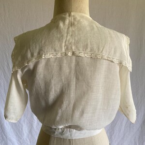 May include: A vintage cream-colored blouse with short sleeves and a high collar. The collar is trimmed with delicate lace. The blouse is displayed on a mannequin, showcasing its tailored fit and classic design. The fabric appears to be lightweight and breathable.