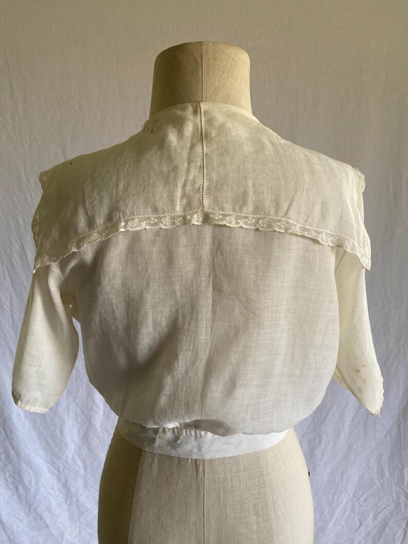 May include: A vintage cream-colored blouse with short sleeves and a high collar. The collar is trimmed with delicate lace. The blouse is displayed on a mannequin, showcasing its tailored fit and classic design. The fabric appears to be lightweight and breathable.