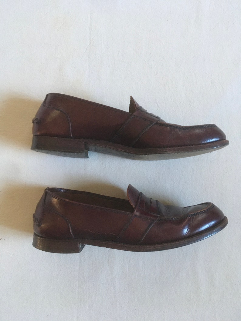 May include: A pair of brown leather loafers with a single strap across the vamp. The loafers have a slightly pointed toe and a low heel.