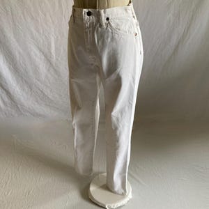 May include: White denim jeans on a mannequin. The jeans feature a classic five-pocket design, a button and zip closure, and a straight leg fit. The fabric appears to be a medium-weight denim. The jeans are displayed on a mannequin.