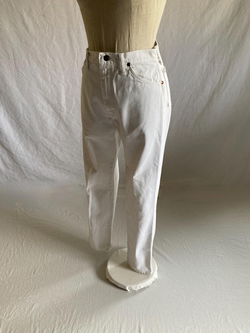 May include: White denim jeans on a mannequin. The jeans feature a classic five-pocket design, a button and zip closure, and a straight leg fit. The fabric appears to be a medium-weight denim. The jeans are displayed on a mannequin.