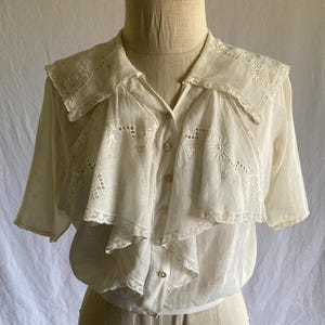May include: Cream-colored vintage blouse with short sleeves and a ruffled collar. The blouse features intricate embroidered floral and geometric patterns. The collar and front panel have delicate lace trim. The blouse is buttoned down the front and is displayed on a mannequin.
