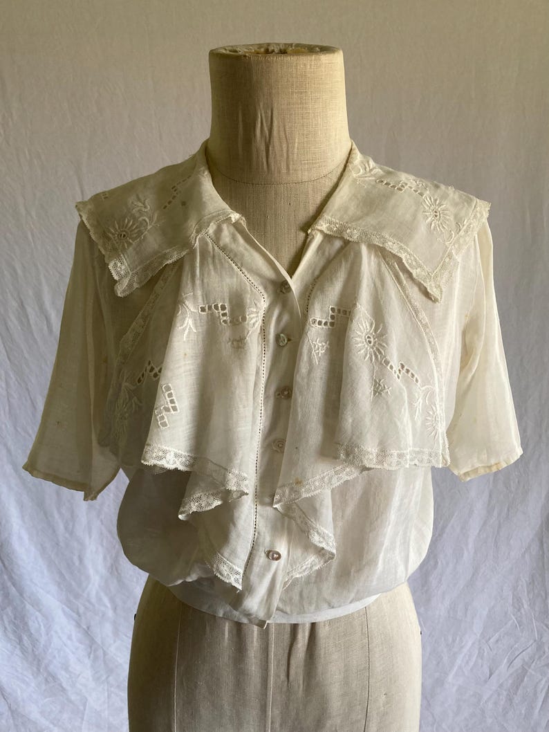 May include: Cream-colored vintage blouse with short sleeves and a ruffled collar. The blouse features intricate embroidered floral and geometric patterns. The collar and front panel have delicate lace trim. The blouse is buttoned down the front and is displayed on a mannequin.