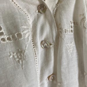 May include: Close-up of a white linen blouse with delicate embroidery and button details. The blouse features a V-neckline and small, pearlescent buttons. Intricate cutwork and floral patterns adorn the fabric, adding a touch of elegance to the garment.