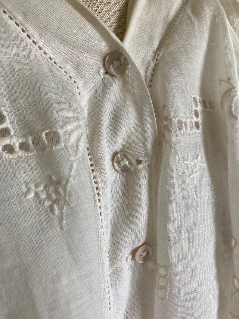 May include: Close-up of a white linen blouse with delicate embroidery and button details. The blouse features a V-neckline and small, pearlescent buttons. Intricate cutwork and floral patterns adorn the fabric, adding a touch of elegance to the garment.