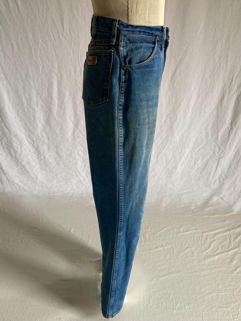 Pu&ograve; includere: Jeans di denim blu chiaro con un classico design a cinque tasche. I jeans hanno un taglio dritto e un lavaggio sbiadito. Una piccola toppa rettangolare &egrave; visibile sulla tasca posteriore. I jeans sono esposti su un manichino.