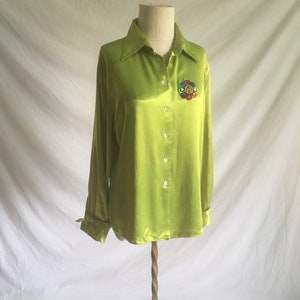 Könnte beinhalten: Ein limegrünes Satinhemd mit langen Ärmeln und Knopfleiste. Das Hemd hat eine bunte gestickte Blume auf der linken Brustseite.